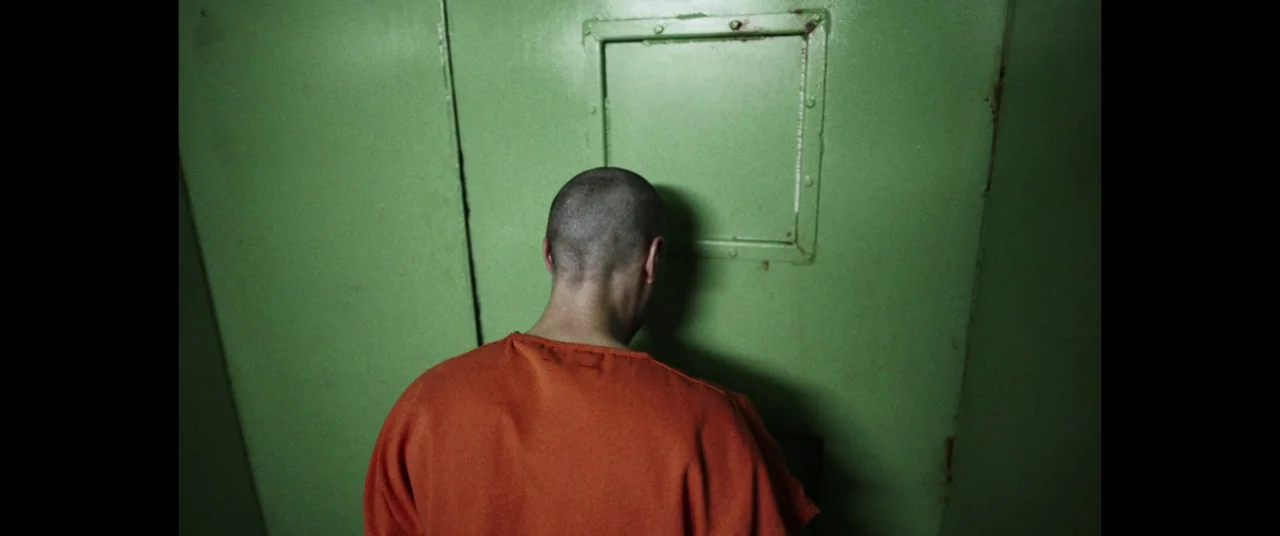 The Mauritanian (2021) movie still: medium — A man in orange scrubs stands before a green metal door with a small window. He leans his forehead…