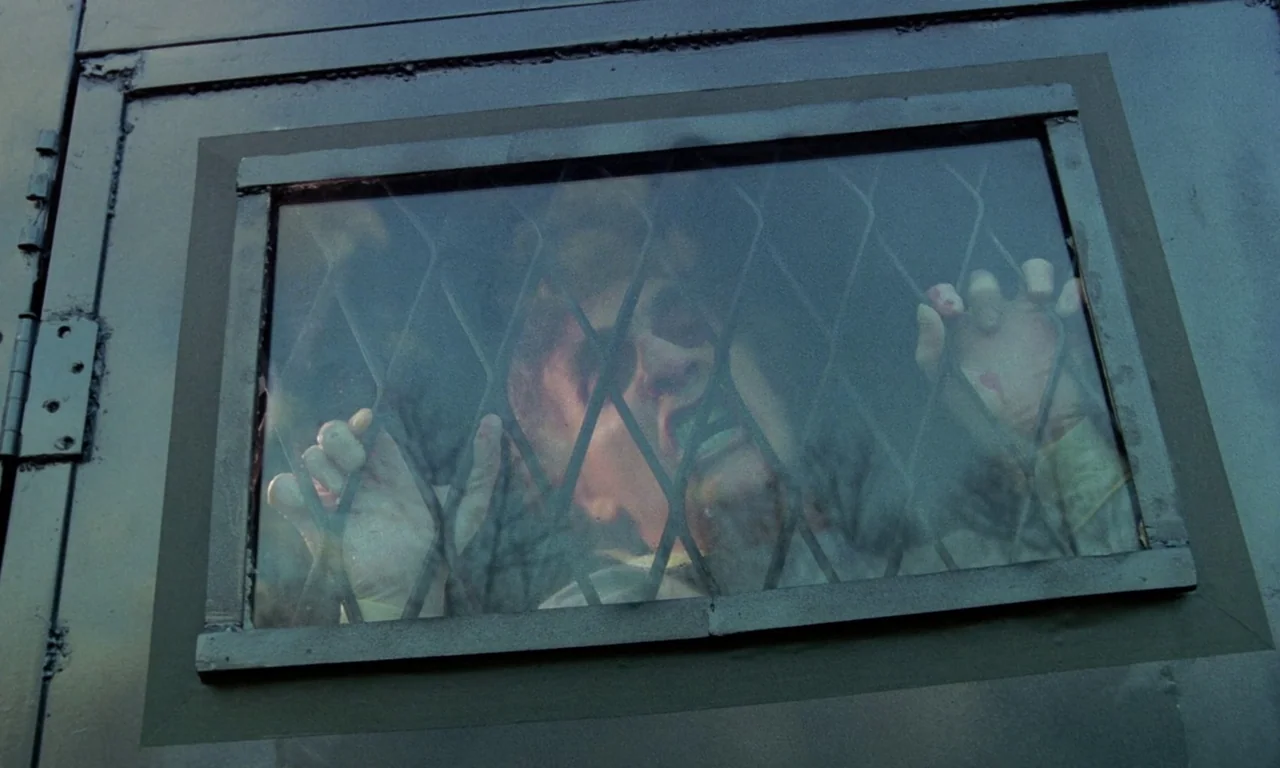 Rabid (1977) movie still: closeup — A dark metal panel features a rectangular viewing portal reinforced with diagonal wire mesh. A man presses his…