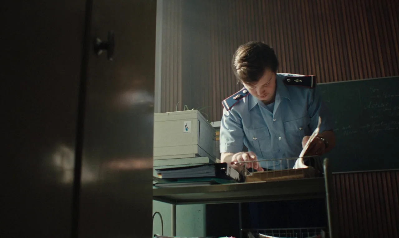 Maldoror (2024) movie still: medium — A workspace with vertical wood paneling and a chalkboard in the background. A man in a blue uniform leans over…