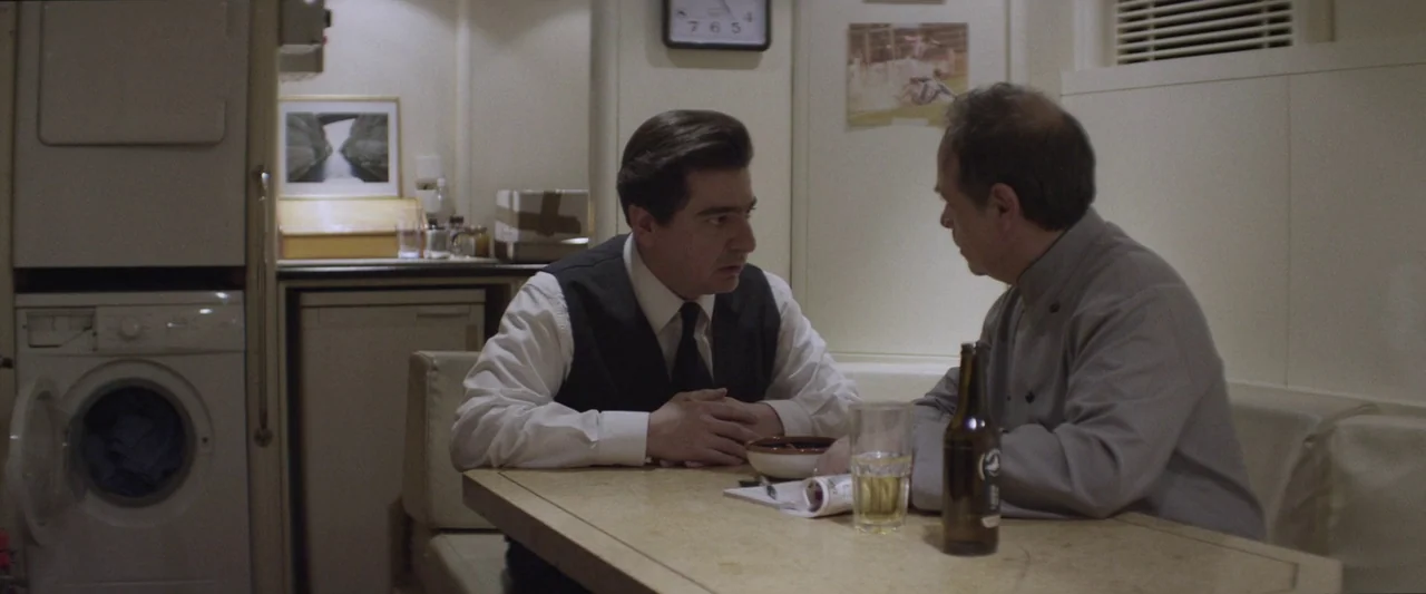 Chevalier (2015) movie still: medium — A small room features a white washing machine, a square wall clock, and photos. Two men sit at a small table,…