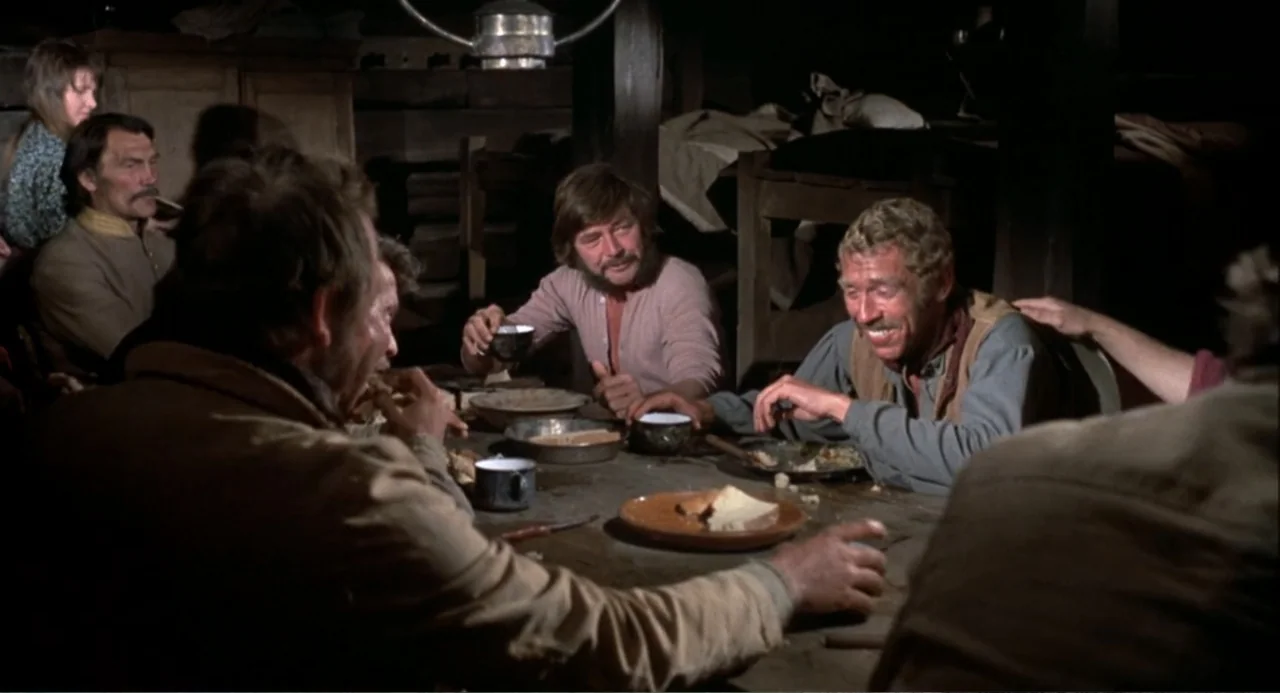 Chato's Land (1972) movie still: medium — A dark rustic room features men gathered around a long worn wooden table. Several men are engaged in…