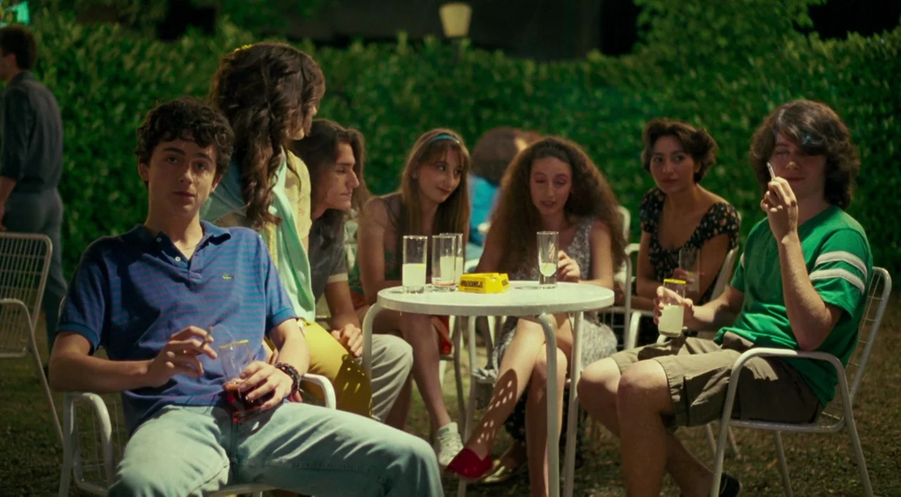 Call Me By Your Name (2017) movie still: medium — A group of young people gather at white metal tables on a grass patio at night. They are seated in…