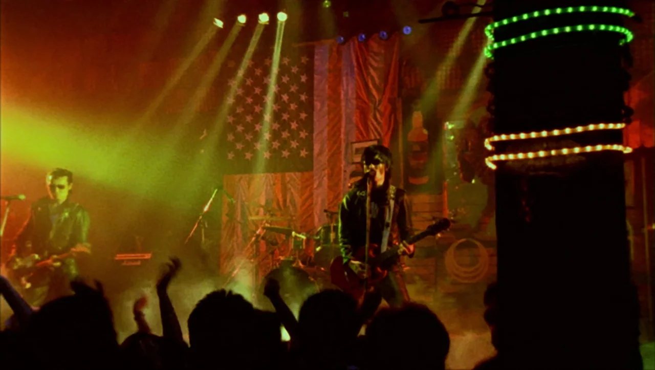 Wild Zero (1999) movie still: wide — A dark, smoky club stage features a band playing instruments in front of an American flag. Silhouetted figures…