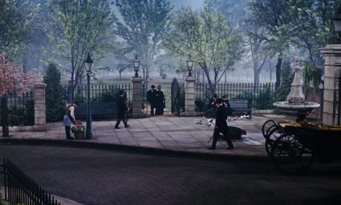Mary Poppins (1964) movie still: wide — A stone-paved entrance to a park features ornate iron fencing and Victorian style lamps. Several people in…