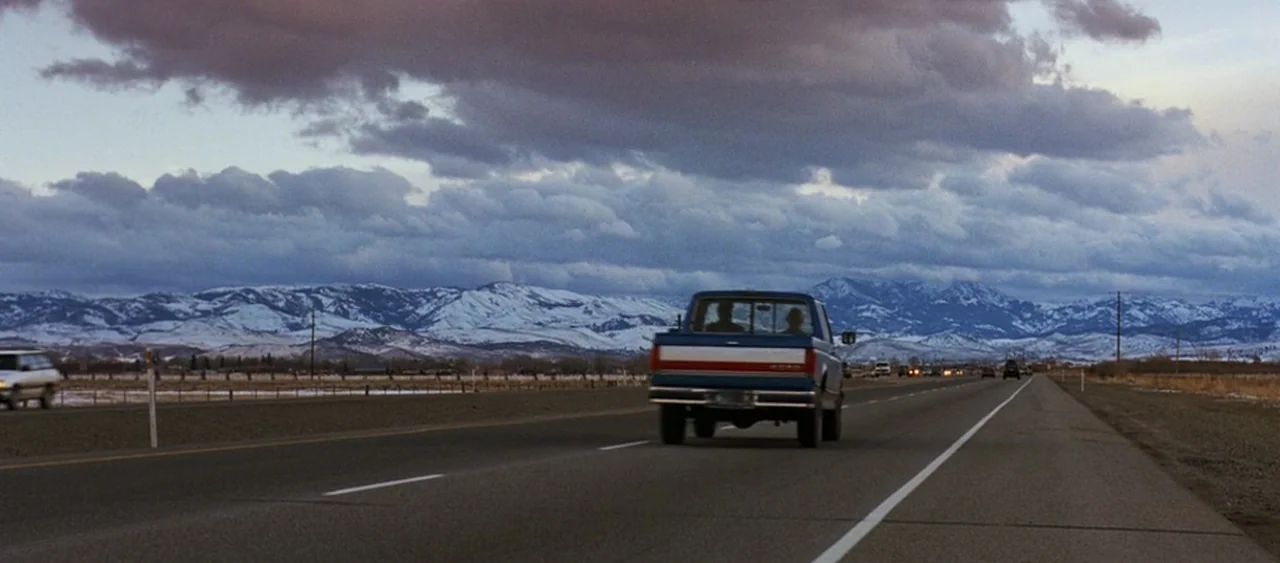 Showgirls (1995) movie still: wide — A dark blue pickup truck drives along a straight highway under a cloudy sky. Snow-capped mountains stretch…