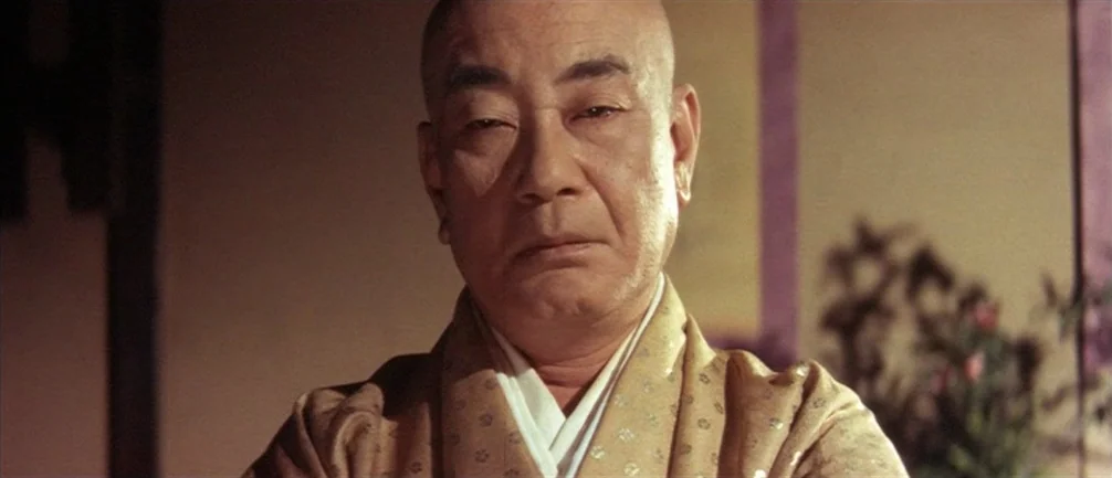 An Actor’s Revenge (1963) movie still: closeup — An interior space with beige walls and a hint of a blurred plant. A middle-aged man with a shaved…