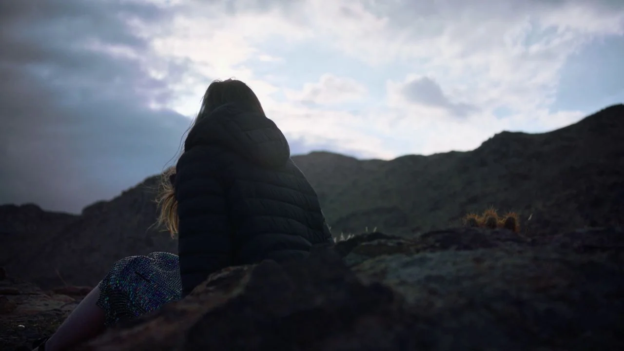 She Dies Tomorrow (2020) movie still: medium — A person sits on a dark rocky slope beneath a cloudy twilight sky. The silhouette of the person…
