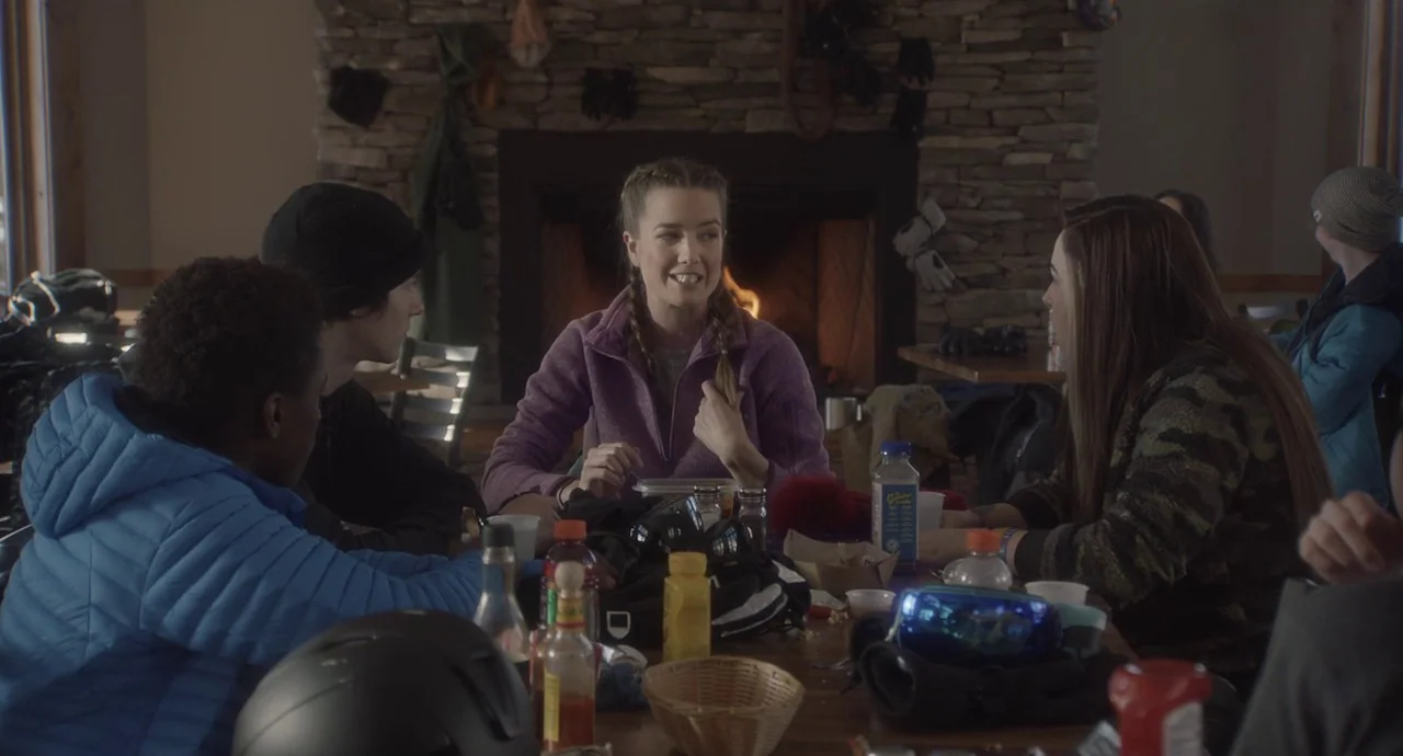 The Wolf of Snow Hollow (2020) movie still: medium — A group of young people sit around a wooden table in a ski lodge. A stone fireplace glows in…