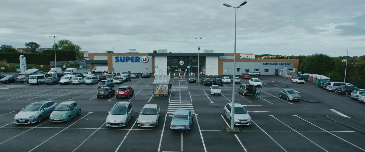 Vincent Must Die (2023) movie still: establishing — A wide exterior shot shows a Super U supermarket with a large asphalt parking lot. Numerous cars…