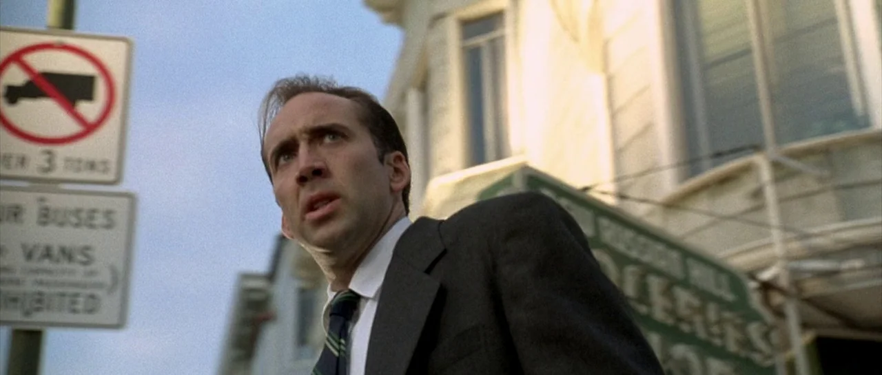 The Rock (1996) movie still: medium — A city street corner features a metal traffic sign and a blurred building facade. A man in a suit looks toward…