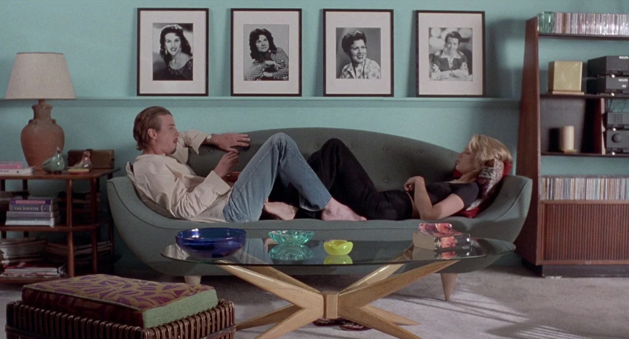 Touch (1997) movie still: wide — A light blue-walled room features mid-century modern furniture, including a curved green sofa. A man and a woman…