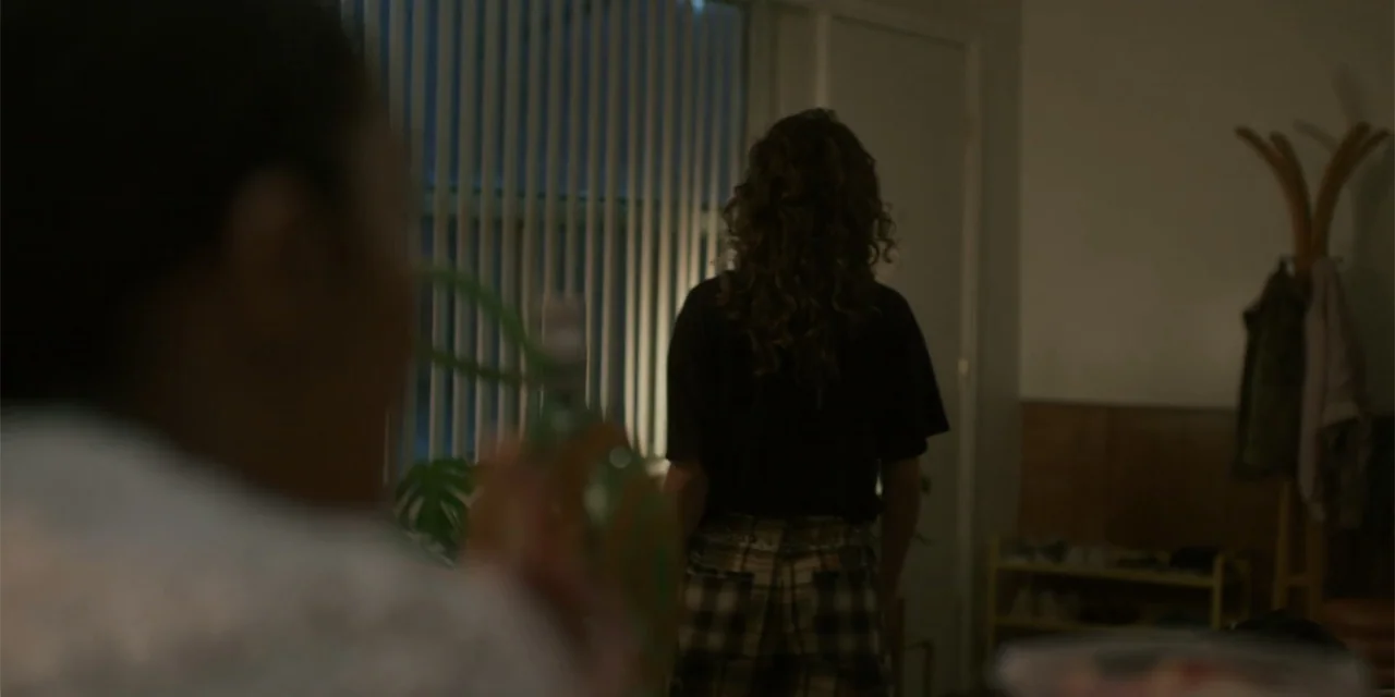 Sitting in Bars with Cake (2023) movie still: medium — A dimly lit room at night features a window covered by vertical plastic blinds. A young woman…