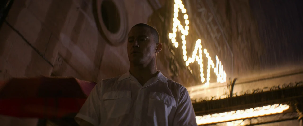 Magic Mike's Last Dance (2023) movie still: medium — An exterior movie theater entrance at night with illuminated marquee bulbs in the rain. A man…