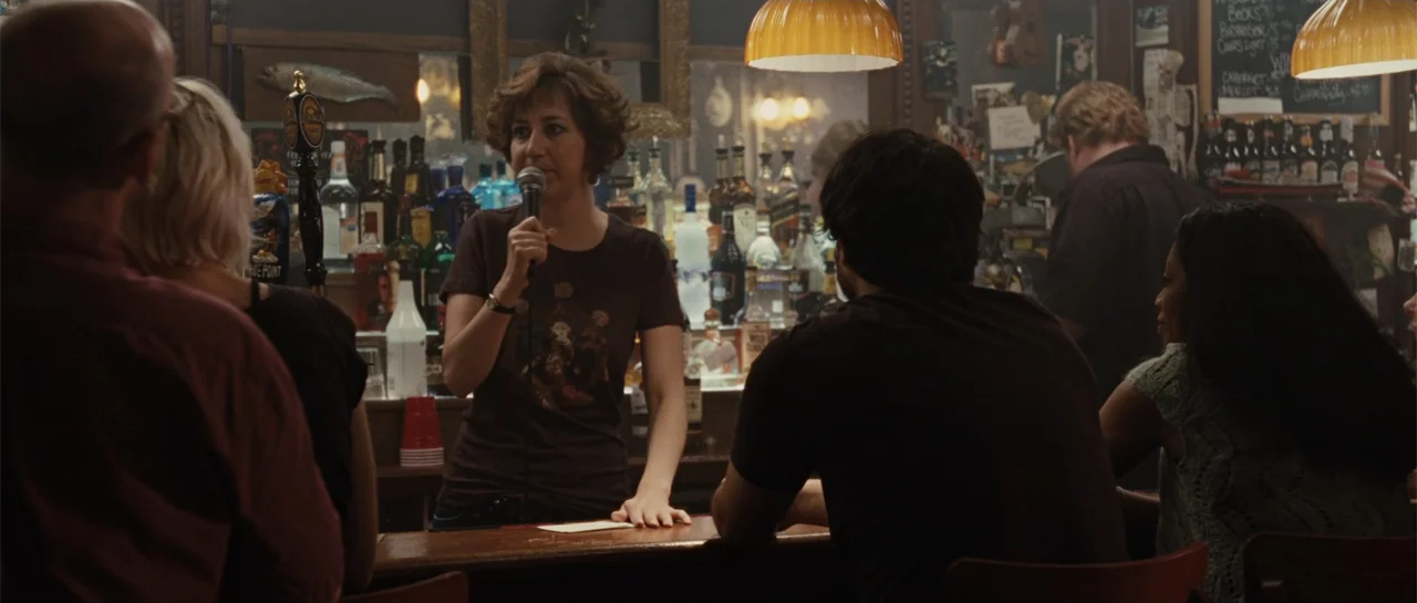 Going the Distance (2010) movie still: medium — A dimly lit bar with numerous bottles stocked on shelves against a dark wall. A woman stands behind…