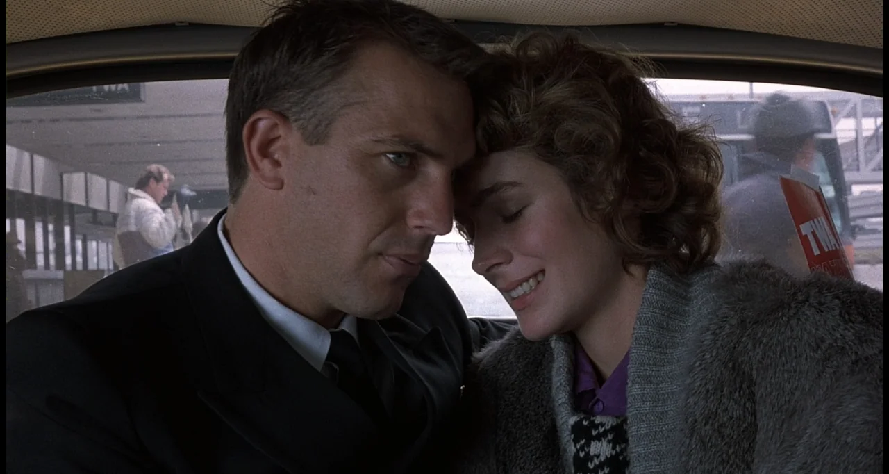 No Way Out (1987) movie still: medium — A man and a woman are seated inside a car parked near a terminal. The pair lean their heads together with…