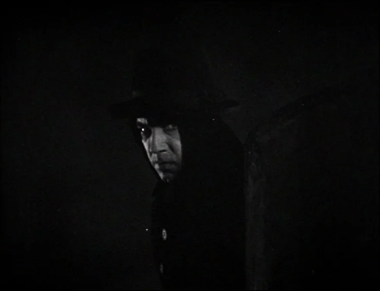 Dracula (1931) movie still: closeup — A dark space features a single vertical architectural edge defining the foreground. A man in a brimmed hat…