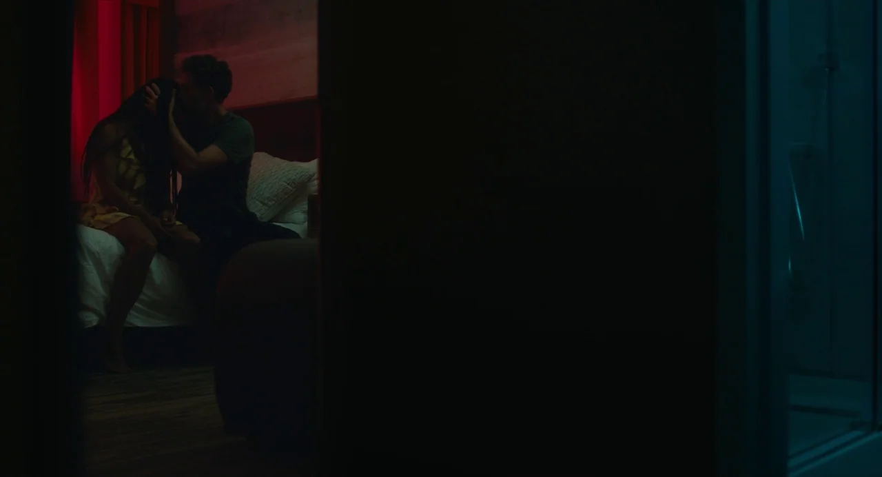 Test Pattern (2019) movie still: wide — A dimly lit hotel room features a bed positioned against a dark wall. A man sits facing a woman while…