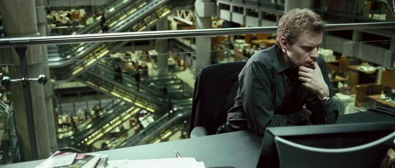 Green Street (2005) movie still: medium — A modern open-plan office space with glass railings overlooks a large atrium. A man sits at a desk in the…
