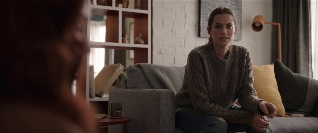 M3gan (2022) movie still: medium — A residential room features a white-painted brick wall and open white shelving units. A young woman sits on a…