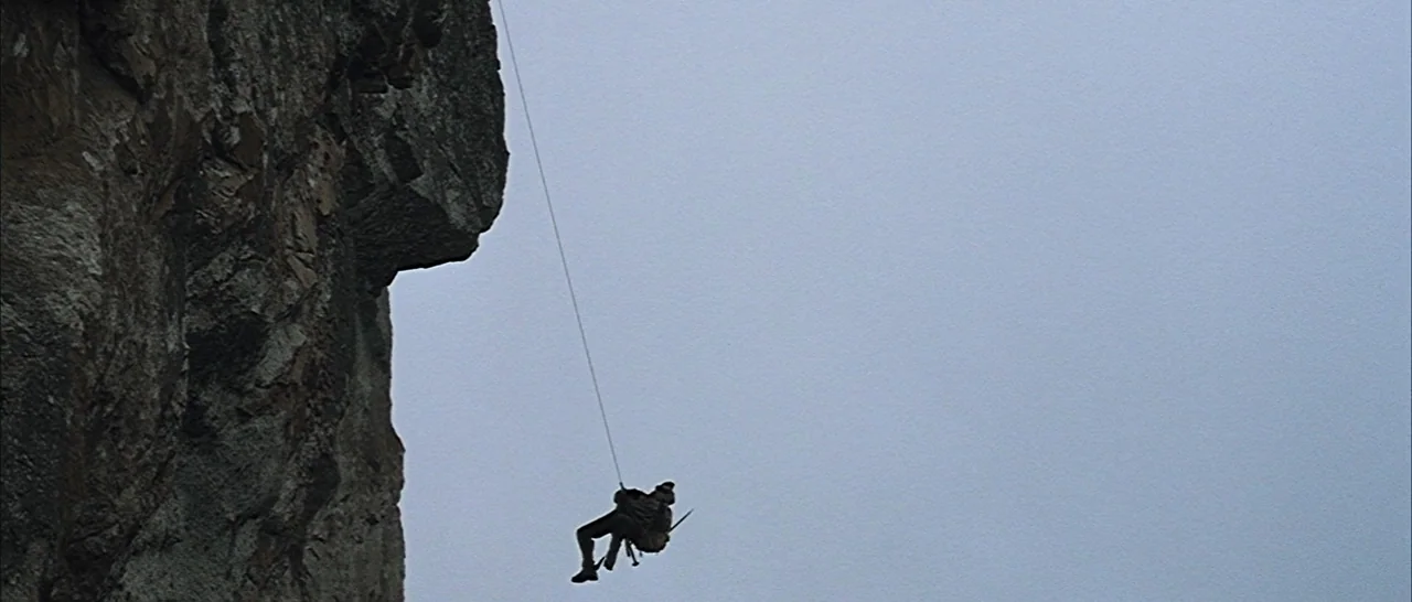 Nymphomaniac Vol 2 (2013) movie still: wide — A vertical gray rock face rises against a vast, overcast sky. A lone person in tactical gear descends…