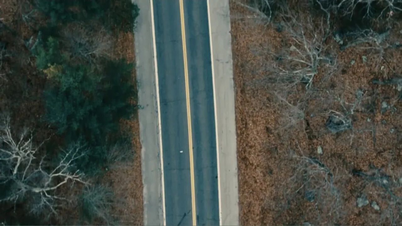 Bleed for This (2016) movie still: establishing — An asphalt road with double yellow lines bisects a landscape of bare autumn trees. The camera…