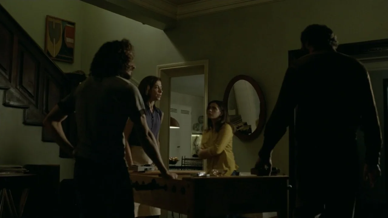 I'm Still Here (2024) movie still: fullbody — A dimly lit room features a wooden foosball table as a focal point. Four adults stand around the table…