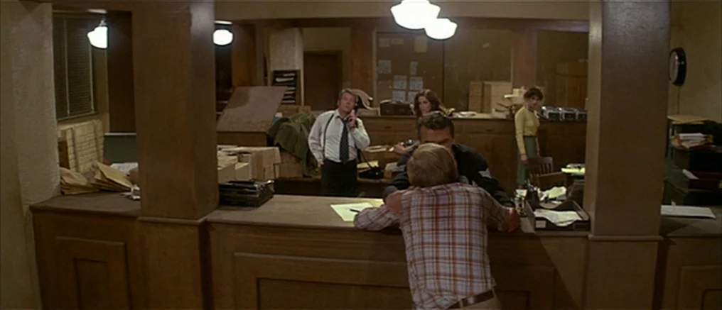 Assault on Precinct 13 (1976) movie still: medium — A busy station interior with wooden counters, scattered paperwork, and several people working…