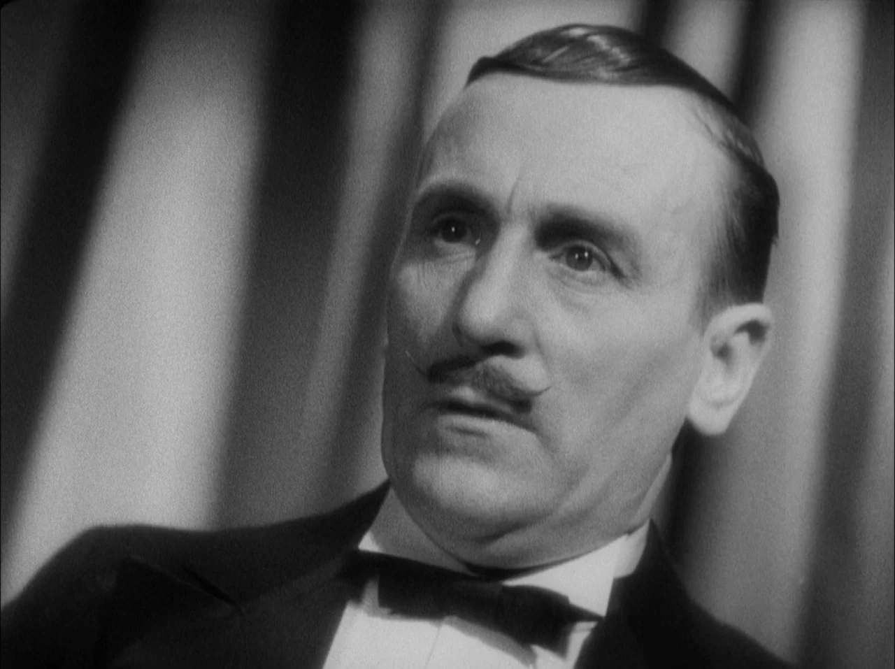 The 39 Steps (1935) movie still: closeup — A man in a tuxedo stands against a backdrop of draped vertical curtains. He looks slightly off-camera…