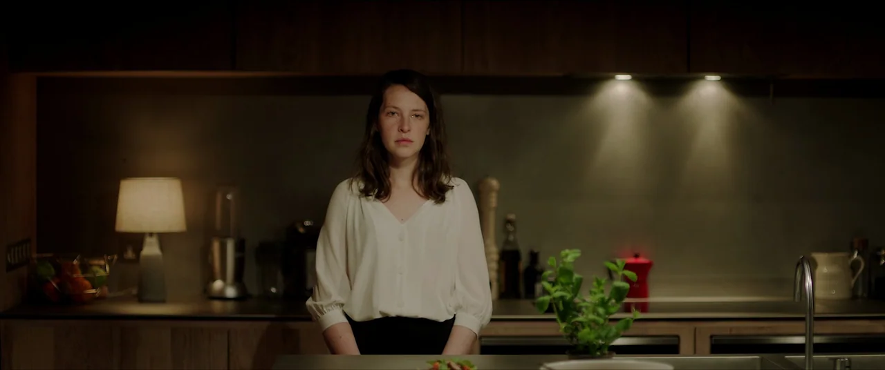 The Feast (2021) movie still: medium — A dark modern kitchen features wood cabinets and minimalist counters illuminated by warm lamps. A woman in a…