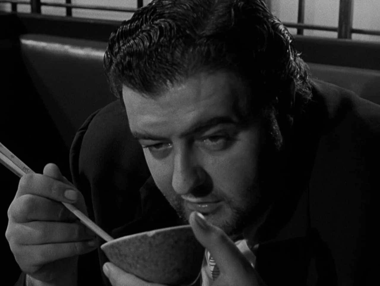 Pickup on South Street (1953) movie still: closeup — A man sits at a dark booth with a wooden slatted divider behind him. He holds a small bowl with…