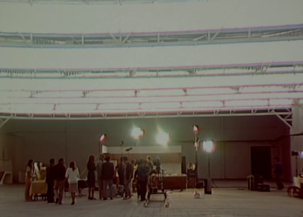 No (2012) movie still: wide — A large, sparsely furnished industrial studio hall features a vaulted metal roof structure above. A film crew with…
