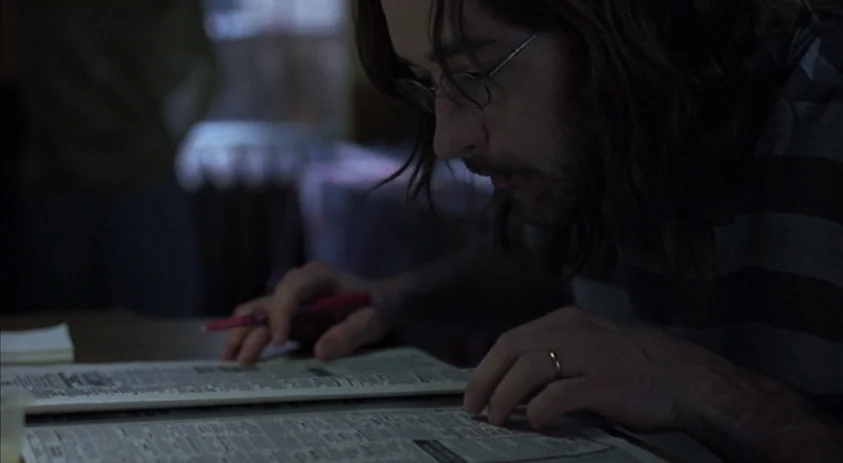Being John Malkovich (1999) movie still: closeup — A dimly lit room contains a man looking closely at a newspaper. He uses a red pen to track lines…