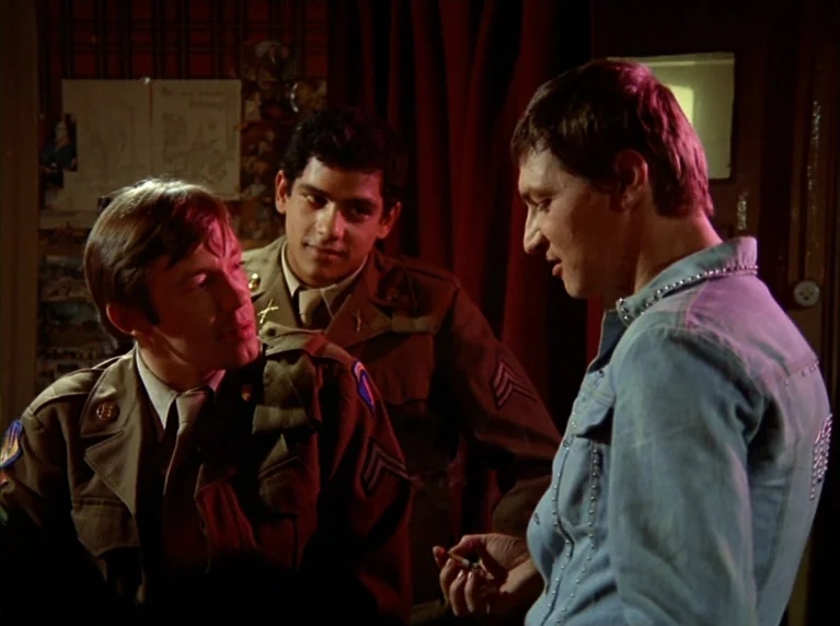 Fox and His Friends (1975) movie still: medium — A dimly lit indoor space features plaid wallpaper and a dark red curtain backdrop. Two men in…
