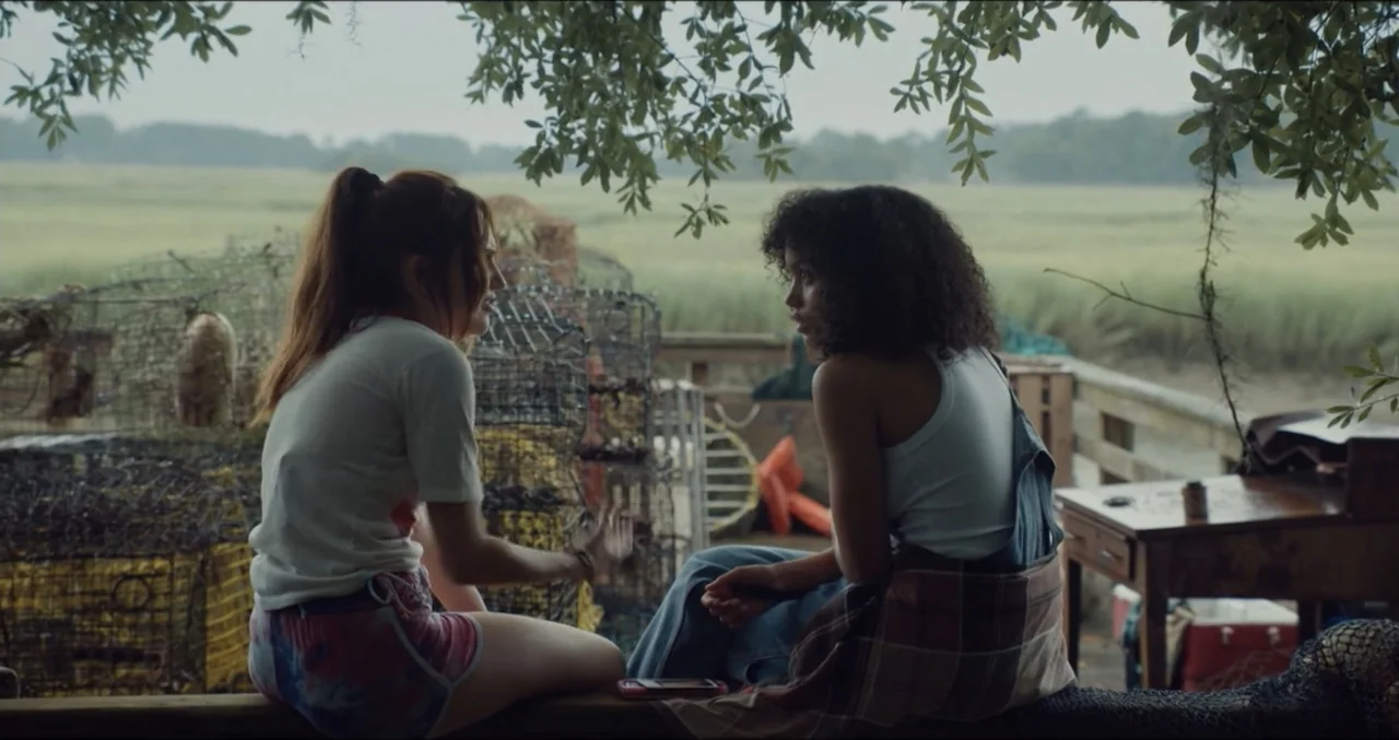 Eye for an Eye (2025) movie still: medium — An outdoor wooden structure overlooks a vast grassy marshland under an overcast sky. Two young women sit…