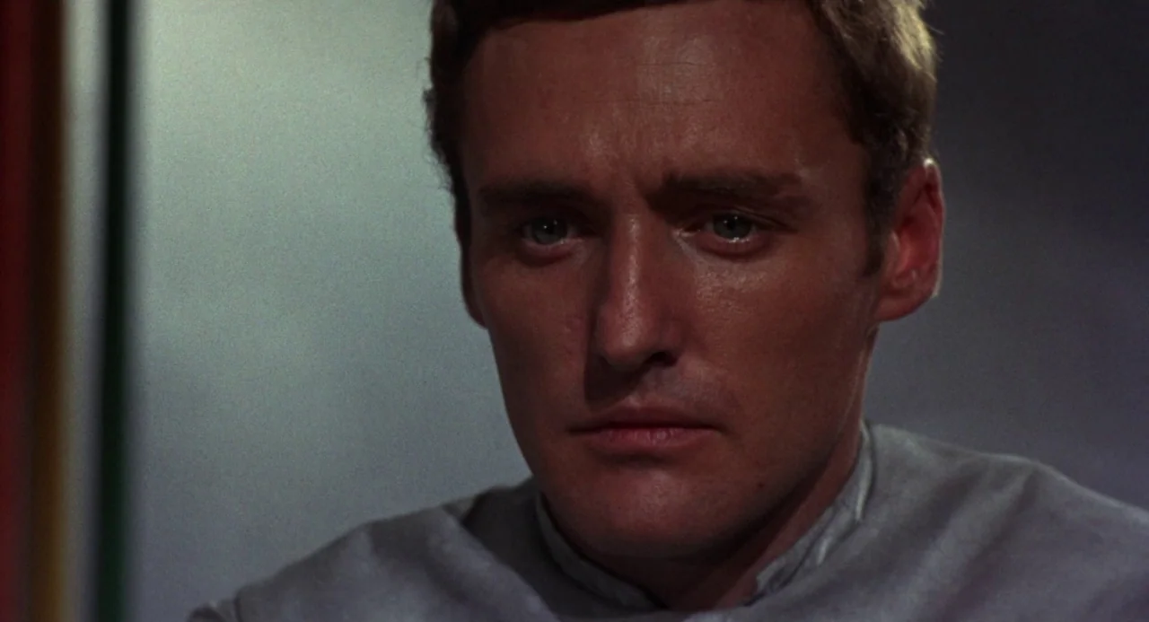 Queen of Blood (1966) movie still: closeup — The frame shows a person in a plain indoor space with dark walls. A man with brown hair and a grey…