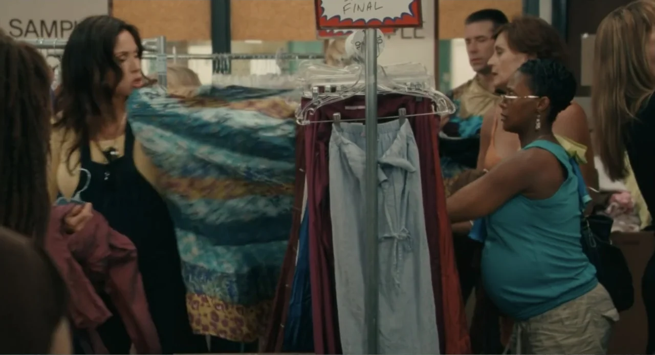 Motherhood (2020) movie still: medium — A crowded retail shop interior features metal clothing racks filled with assorted fabric garments. A woman…