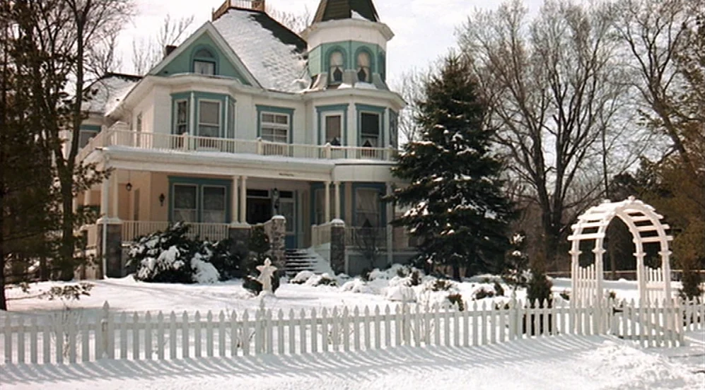 Groundhog Day (1993) movie still: wide — A two-story Victorian house with white siding and teal trim sits behind a fence. The lawn and trees are…