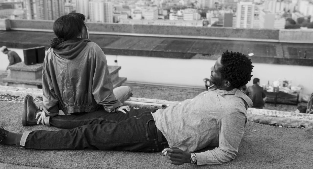 Paris, 13th District (2021) movie still: medium — A rooftop surface covered in artificial grass overlooks a dense urban skyline. Two people lounge…