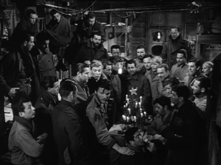 Stalag 17 (1953) movie still: wide — A dimly lit room crowded with soldiers in winter military attire. They gather around a small table displaying a…