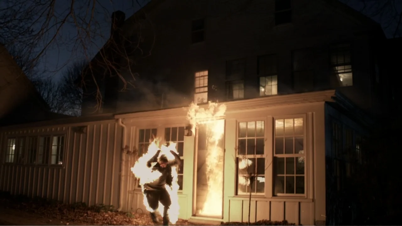 The Collective (2023) movie still: wide — A two-story residential house at night has a burning entrance with flames rising vertically. A person…