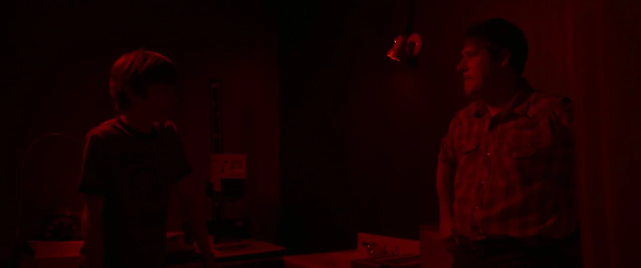 Summer of 84 (2018) movie still: medium — A darkroom lit exclusively by a red safe light contains a photo enlarger machine. A man and a boy stand…