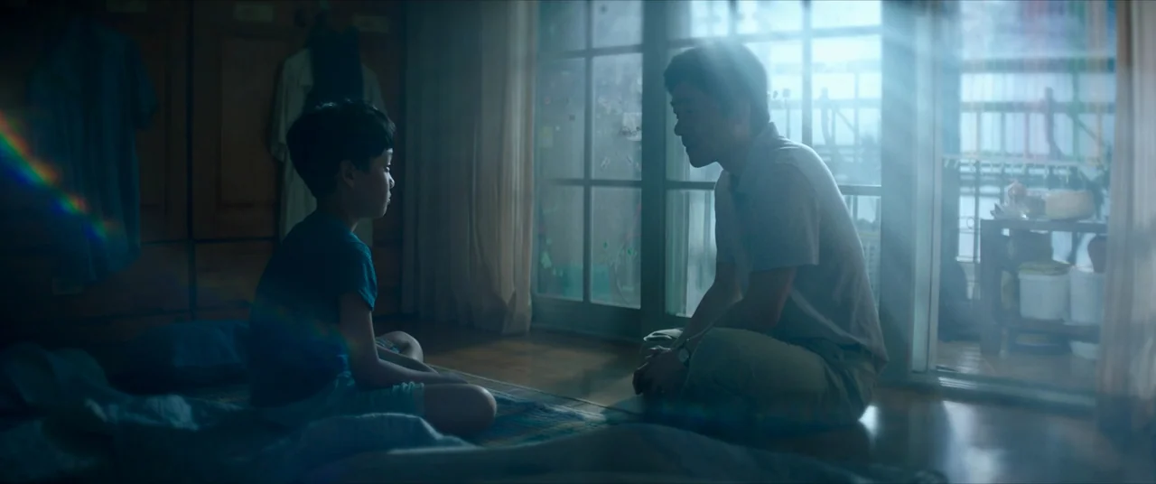The Divine Fury (2019) movie still: medium — A cluttered room features dark wood paneling and large sliding glass doors leading outside. A man and a…