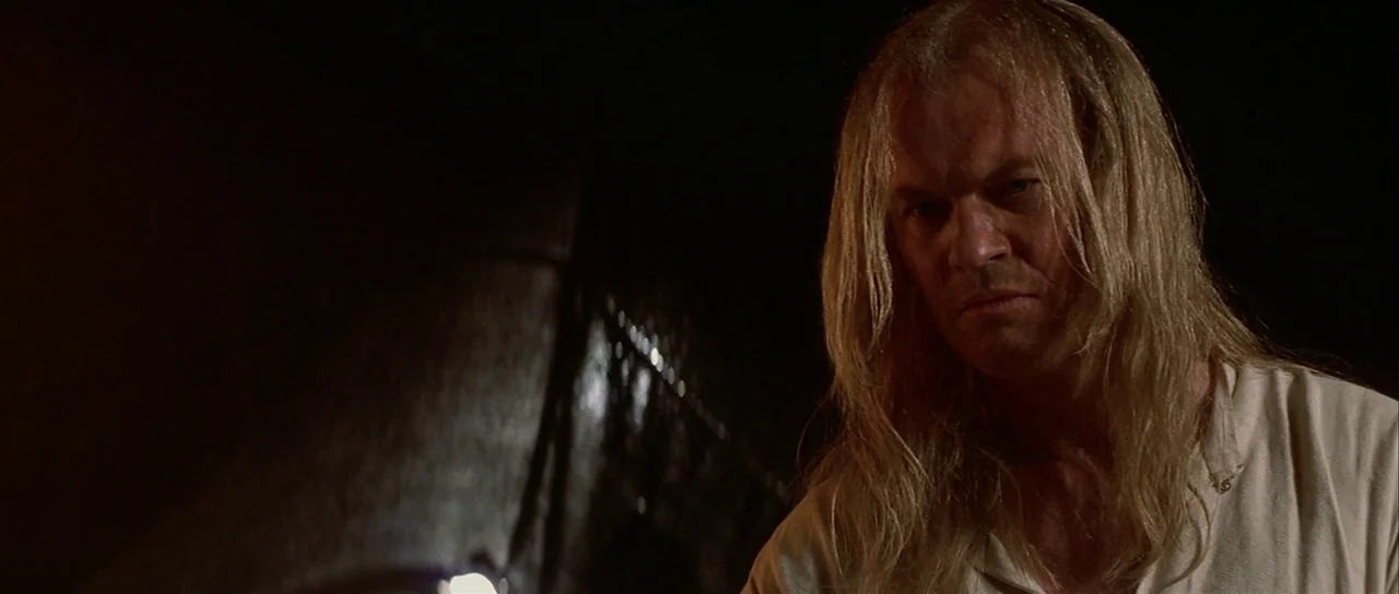 The 13th Warrior (1999) movie still: medium — A man stands in a dark, indistinct interior space with deep shadows. The subject gazes intently toward…