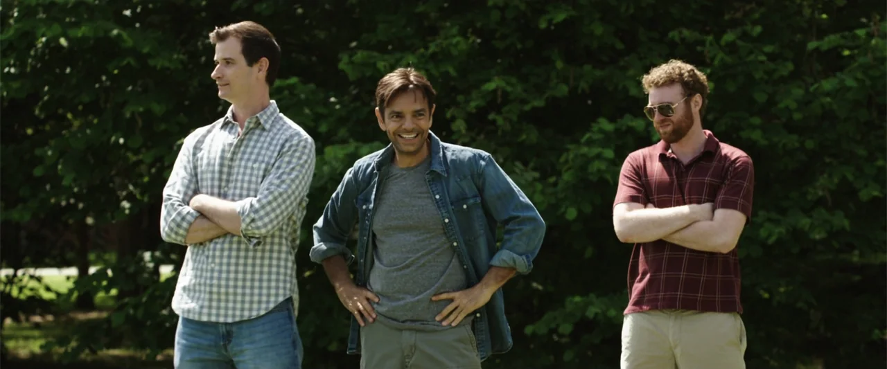 Overboard (2018) movie still: fullbody — Three men stand in a park with dense green trees visible in the background. They are positioned…