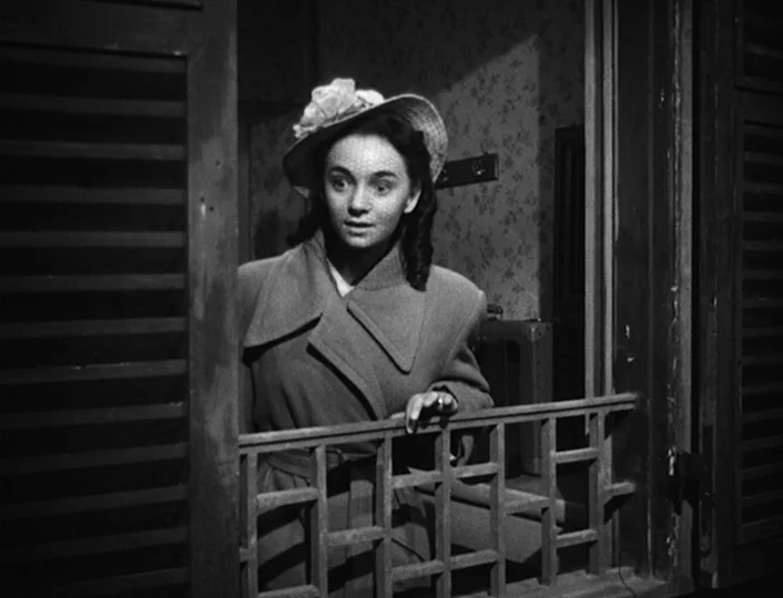 The White Sheik (1952) movie still: medium — A woman stands behind a wooden lattice window frame in a dim room. She leans forward looking out from…