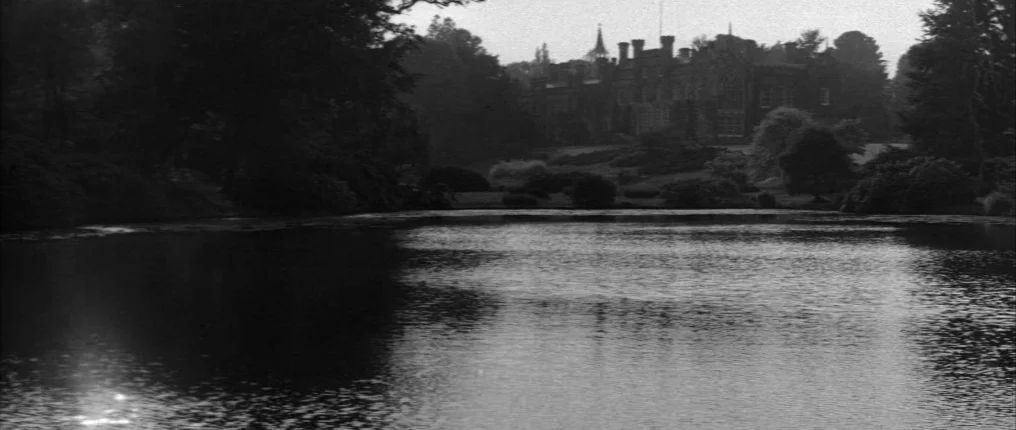 The Innocents (1961) movie still: establishing — A dark, still lake foregrounds a large stone mansion nestled among dense, mature trees. The…