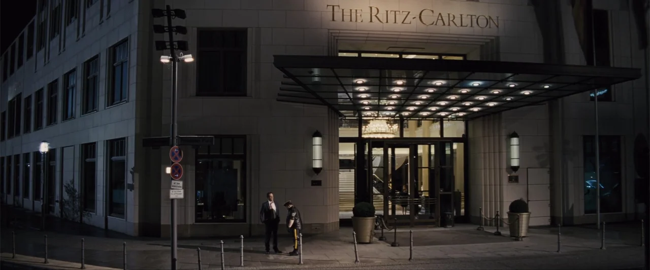 Unfinished Business (2015) movie still: wide — The exterior of a luxury hotel at night with gold lettering above the entrance. Two men stand on the…