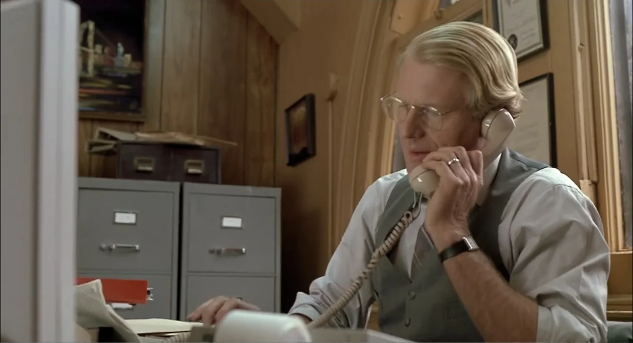 She-Devil (1989) movie still: medium — An office with grey metal filing cabinets and wood paneling on the wall. A middle-aged man in a dress shirt…