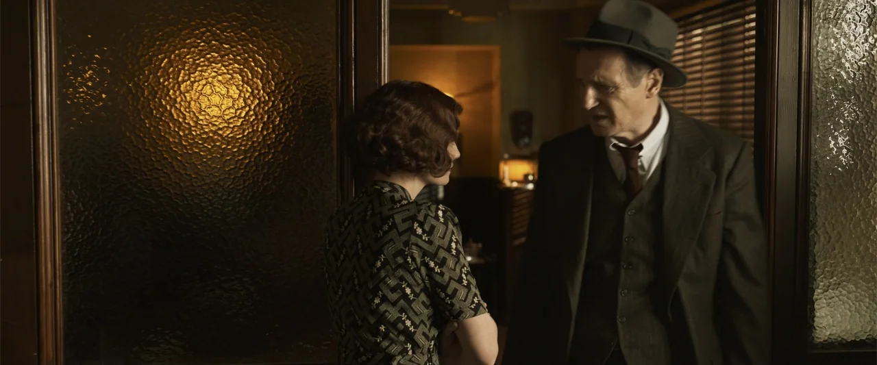 Marlowe (2023) movie still: medium — A dark, wood-paneled interior room features textured glass panes in a doorway. A man in a suit and hat stands…