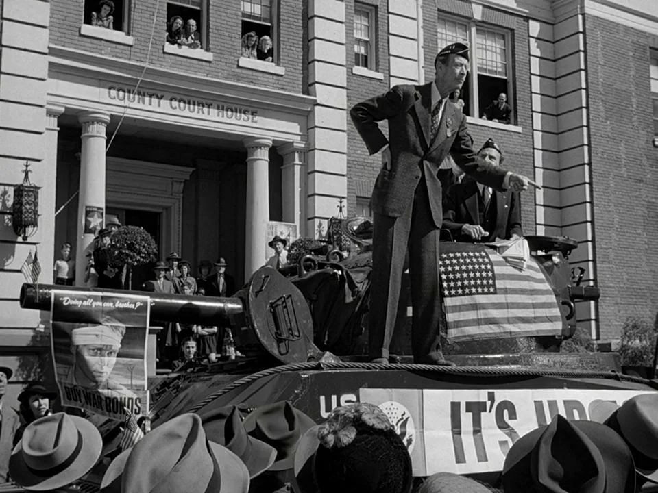 It’s A Wonderful Life (1946) movie still: wide — A stone building with large columns stands behind a crowd gathering outdoors. A man in a suit…